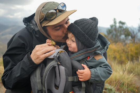Father Kissing Child In Backpack Carrier On Mountain Trail