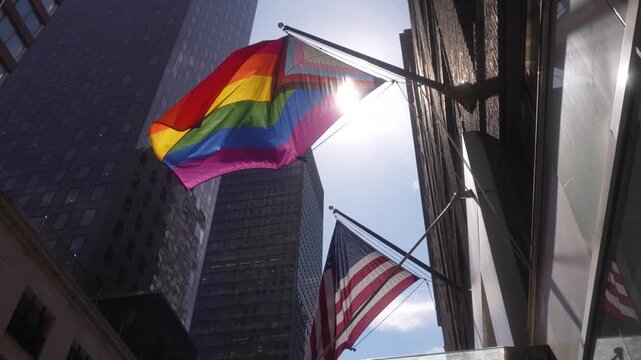 A cinematic video clip of an LGBTQ flag and an American flag waving in front of skyscrapers on a sunny day.
