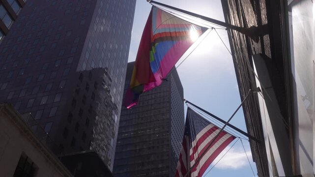 A cinematic video clip of an LGBTQ flag and an American flag waving in front of skyscrapers on a sunny day.
