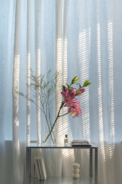 Pink lilies and personal objects on sunlit table by sheer curtains
