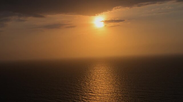Golden sun setting over the calm sea horizon with beautiful reflections on the water and a cloudy sky. A serene seascape showing a vibrant orange glow at dusk