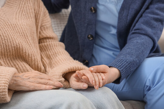 Senior woman with dementia and caregiver holding hands on sofa at home, closeup