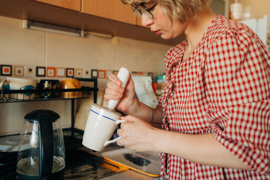 Woman frothing milk in mug near kettle in kitchen