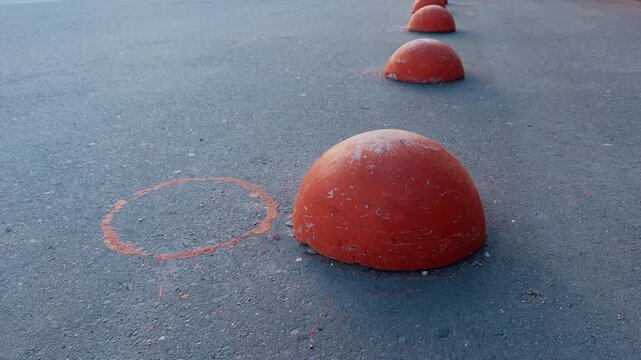 Red hemispheres on asphalt pedestrian path, row of weathered orange bollards creating rhythmic barrier in empty urban pedestrian