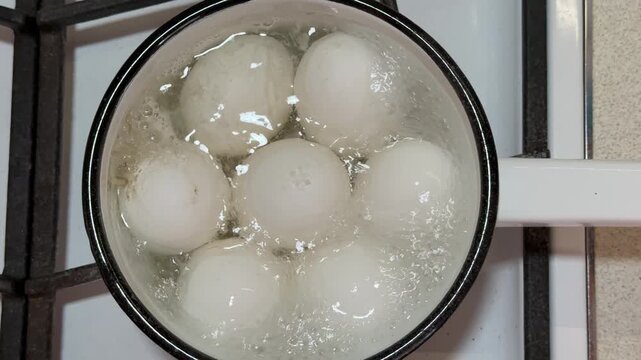 Seven white eggs boiling in ladle over gas stove close-up, enamel ladle filled with bubbling water and rising steam, glossy shells