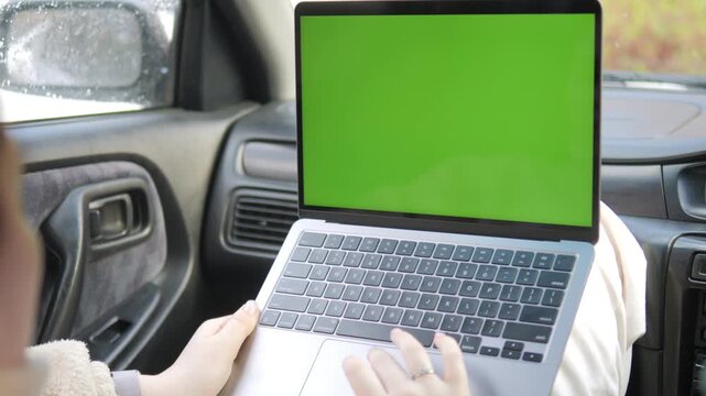Person uses laptop with green screen inside car. Hands type on keyboard. Focus stays on screen for chroma key editing. Suitable for remote work, travel, or digital content. Rain visible on windshield