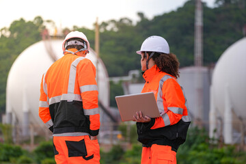 Obraz premium Two engineers in high-visibility safety gear and hard hats discuss project plans on a laptop in front of large spherical gas storage tanks at an industrial coastal facility.