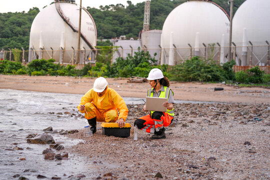 Two engineers in safety gear collect water samples and input data on a laptop near coastal chemical storage tanks. Field research and pollution monitoring in action.