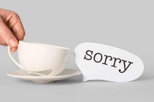 Female hand with broken tea cup and word SORRY on grey background