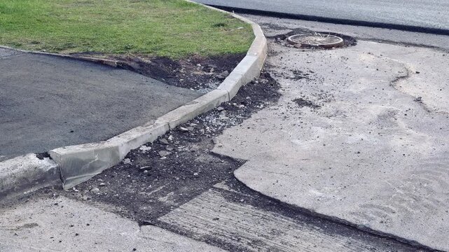 Damaged asphalt curb corner with aggregate, scattered loose asphalt and gravel along cracked pavement next to grassy verge