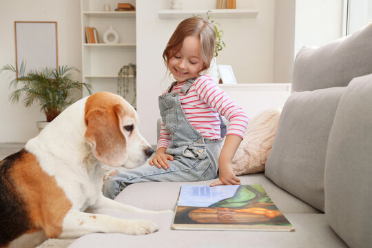 Cute little girl with Beagle dog reading book on sofa in living room