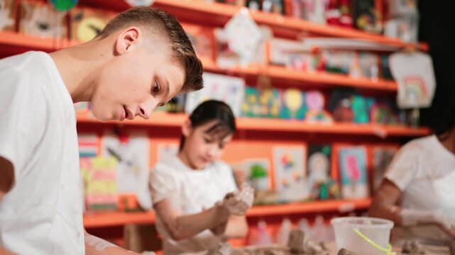 Asian high school student modeling clay on table with mud and bottle of water at pottery workshop. Group of happy diverse children working or making cup of clay. Creative activity concept. Edification