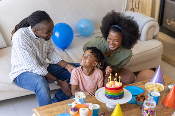 Obraz premium Diverse family celebrating birthday in living room at coffee table with rainbow cake and balloons