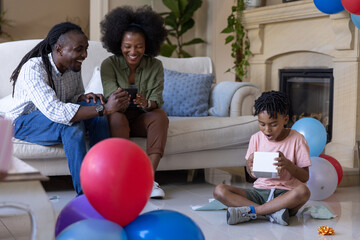 Obraz premium African American family sitting in living room smiling at phone, child opening gift box, copy space