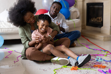 Obraz premium African American family sitting on living room floor, mother hugging child, balloons and confetti