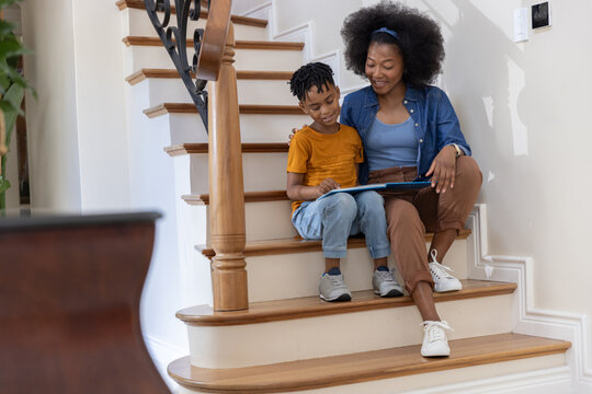 African American mother and child sitting on wooden staircase reading children's book near newel