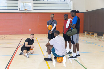 Fototapeta premium Diverse group of five men standing holding basketballs on gym court with towel and duffel bag