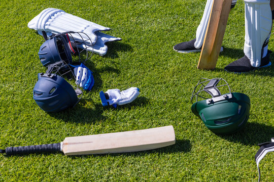 Cricket bat, helmets, gloves and pads are lying scattered on well-trimmed grass under bright sun