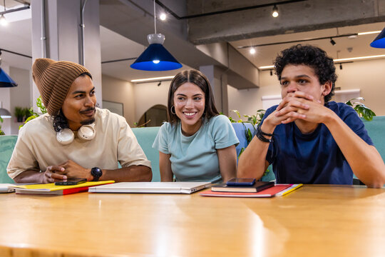 Diverse friends chatting at long wooden table in coworking lounge with closed laptop and notebooks