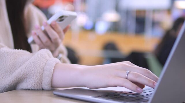 Close-up shows hands typing on laptop while holding phone. Person appears focused, multitasking in casual indoor setting. Suitable for content about remote work, digital communication