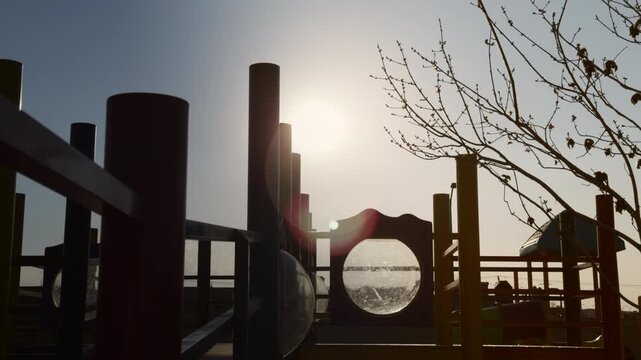 Tranquil silhouette of empty modern outdoor children's playground in public or preschool park with sun glare and tree branches swinging - peaceful, calm, isolation or lonely concept