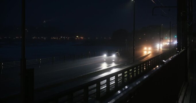 Headlights of vehicles on wet street near ocean in Japan at night during storm