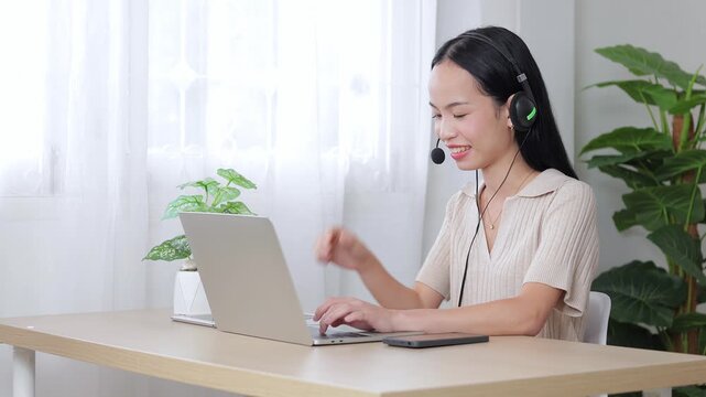 A beautiful smiling Asian woman wearing a headset while working on her laptop from a bright home office, providing friendly customer support, online consulting, or remote teaching services.