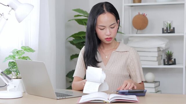 Anxious Asian woman checking financial documents and counting family expenses with a calculator. She looks stressed and concerned about high bills and inflation at her desk.