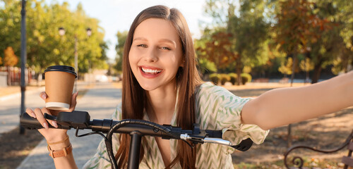 Obraz premium Beautiful young happy woman with cup of coffee riding electric kick scooter in park