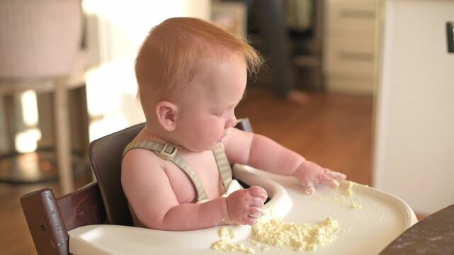 Baby with light red hair in high chair engages with food, covering hands and tray with mess, while a caregiver stands in the background, highlighting a joyful and interactive mealtime experience