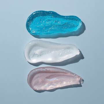 Three different cosmetic textures including a translucent blue gel, an opaque white cream, and a smooth pink lotion are smeared horizontally on a soft blue background.