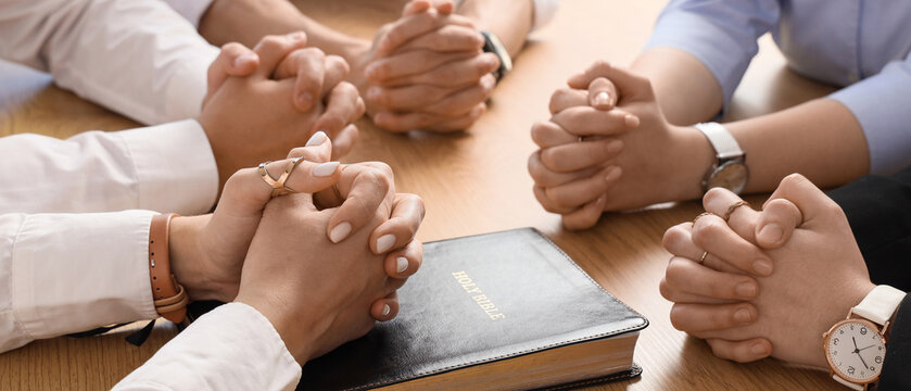 Group of people praying with Holy Bible at table in office, closeup