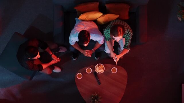 Happy family and colleague sitting at dark living room with red filter while cheering football team together. Caucasian people enjoy watching tv and giving high five and clinking glasses. Convocation.