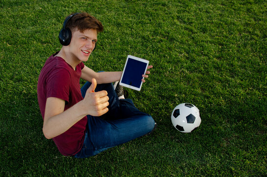 Blonde teenager sitting on the fresh cut grass holding tablet with empty screen and soccer ball next to him, showing thumbs up to camera