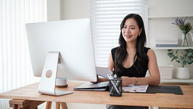 Smiling businesswoman working at a desktop computer while reviewing documents in a bright modern office.