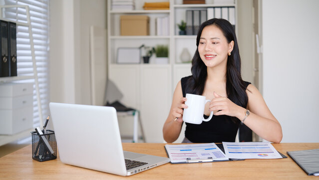 Professional businesswoman enjoying a coffee break while reviewing business charts and documents in a bright office.