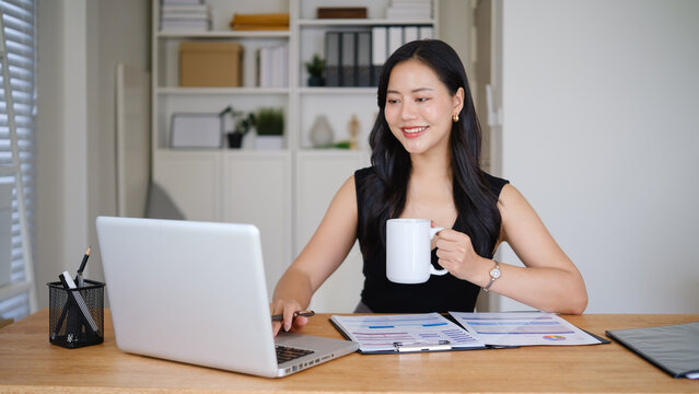 Professional businesswoman enjoying a coffee break while reviewing business charts and documents in a bright office.