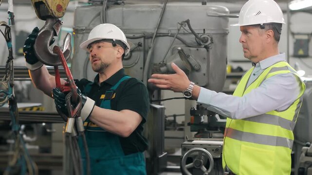 Industrial workers inspecting lifting hooks and rigging equipment inside a manufacturing facility. Workplace safety, machinery maintenance and industrial teamwork in production environment.