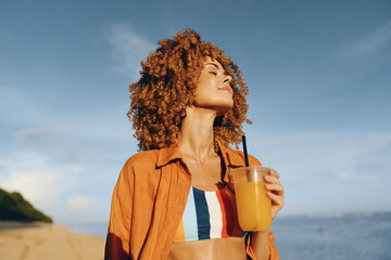 Young woman with curly red hair enjoys a refreshing drink on a sunny beach while wearing a colorful...