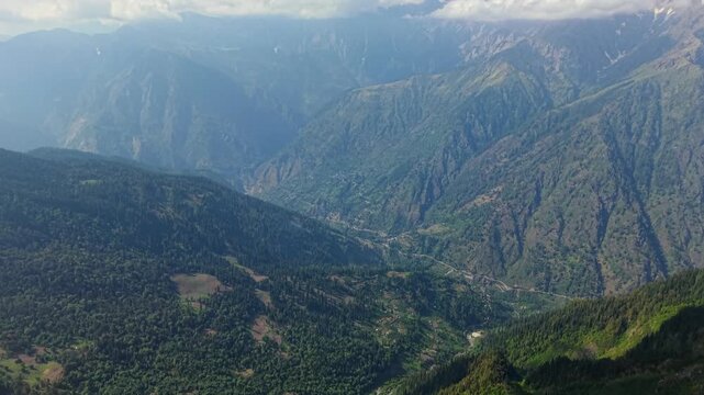 Tilt up drone shot revealing Parvati Valley rising toward snow covered Himalayan peaks beneath clouds.