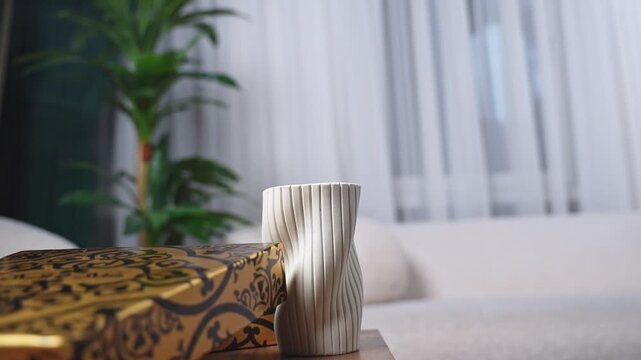 Close-up of rising incense smoke beside a white ribbed ceramic vase and golden patterned box in a minimalist living room. Soft daylight illuminates the peaceful indoor home environment.