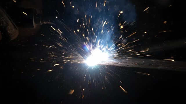 Close-up of industrial metal arc welding process in a dark factory environment featuring bright white light and glowing orange sparks spraying across a steel plate.