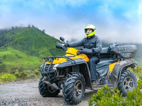 Adventure tourism. Rider drives yellow ATV on muddy mountain trail through green hills and misty landscape.