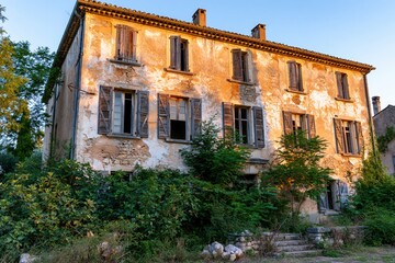 Obraz premium Abandoned weathered French country house with peeling paint and overgrown vegetation in golden hour light.