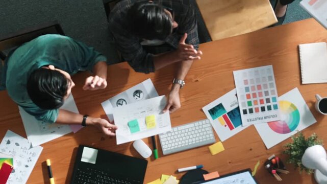 Top view of manager writing idea while team standing and giving comment. Professional designer design logo while looking at color palette and writing idea down in paper with color marker. Symposium.