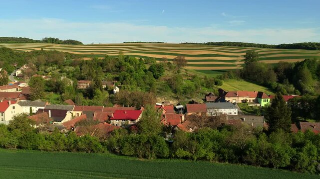 Aerial drone view of a small Czech village with terracotta rooftops bordered by striped crop fields with undulating patterns on surrounding hillsides, Moravia, Czech Republic.