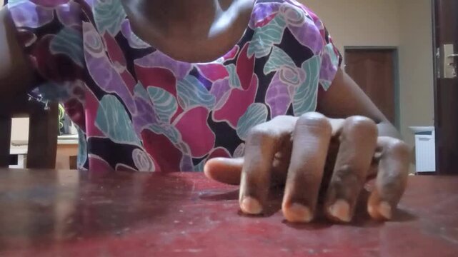 African tapping fingers on brown table. Black woman in floral dress hitting finger on coffee color wooden table at home. Close up of one hand in systematic fast motion, rhythm action.