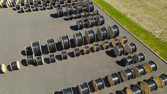 Drone view of numerous wooden and metal spools with black electrical or fiber optic cables stored outdoors in an industrial area on a sunny day, showcasing production and logistics