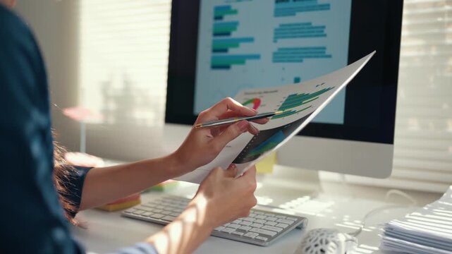 Close up of a female financial analyst comparing data from a document to charts on a computer screen, working at her office desk