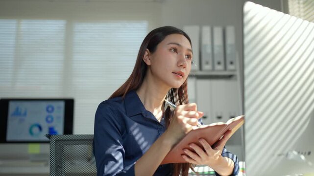 Young professional Asian businesswoman working at her desk, writing important notes in her daily planner and listening during a meeting